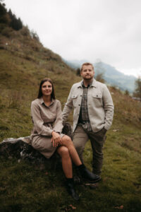 Paar in Outdoor-Kleidung in herbstlicher Berglandschaft, Frau sitzt auf einem Stein, Mann steht daneben